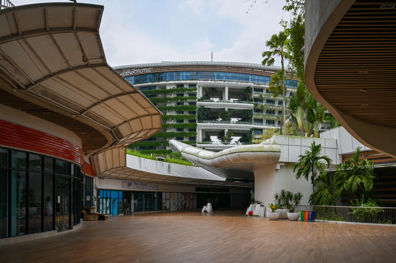 A modern-looking plaza is seen beneath a partially cloudy sky. The shops lining either side of the walkway are closed, dark behind their glass windows, and a hotel building looms in the background, with green plants hanging from its balconies. No people are visible either in the plaza or in the hotel.