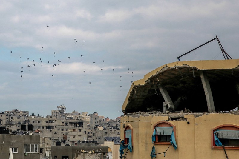 Aid parcels drift through the sky over the battered remains of densely packed urban buildings in the northern Gaza Strip. Each parcel is attached to a parachute, and they appear as small dots against a cloudy gray sky. A building in the foreground has a partially caved-in roof, and the window glass is blown out.