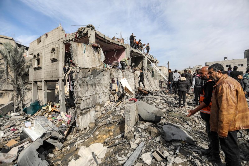 Palestinians stand outside of a mostly destroyed building in the Gazan city of Khan Younis. The two men closest to the camera speak to each other while gesturing at the rubble. Farther in the background, people climb hunks of broken concrete as they ascend to the roof and search for survivors in the wreckage.