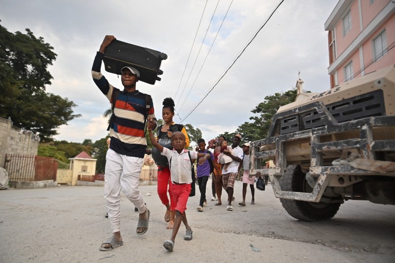 A group of people walk across faded concrete beneath a cloudy sky as they flee gang violence in a neighborhood of Port-au-Prince, Haiti. A man with a suitcase balanced on his head leads the group, using his free hand to hold the hand of a child walking alongside him. A woman with belongings in a trash bag follows, and a man carrying a toddler and other people are visible in the background.