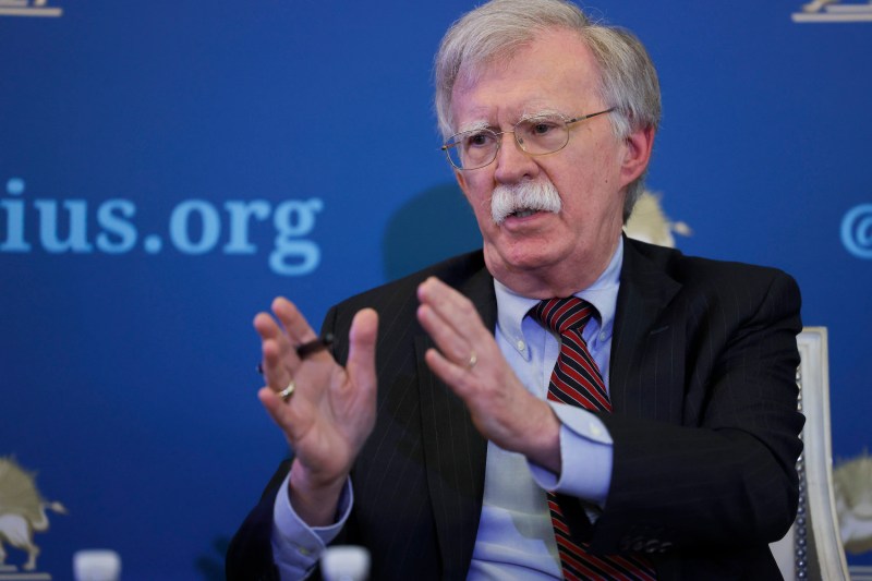 Former U.S. National Security Advisor John Bolton gestures with both hands while he speaks at a panel. Bolton is a man in his 70s wearing a navy blue suit, a striped tie, and wire-framed glasses.