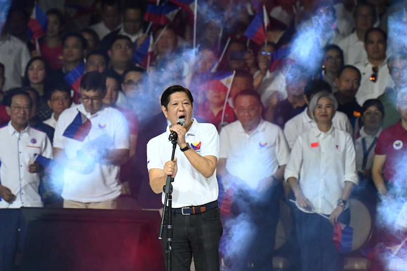 Philippine President Ferdinand Marcos Jr. stands and grips a microphone stand as he delivers a speech during a rally. Rows of supporters stand behind him, and wafts of smoke or steam are visible in shafts of light that hang over the crowd.