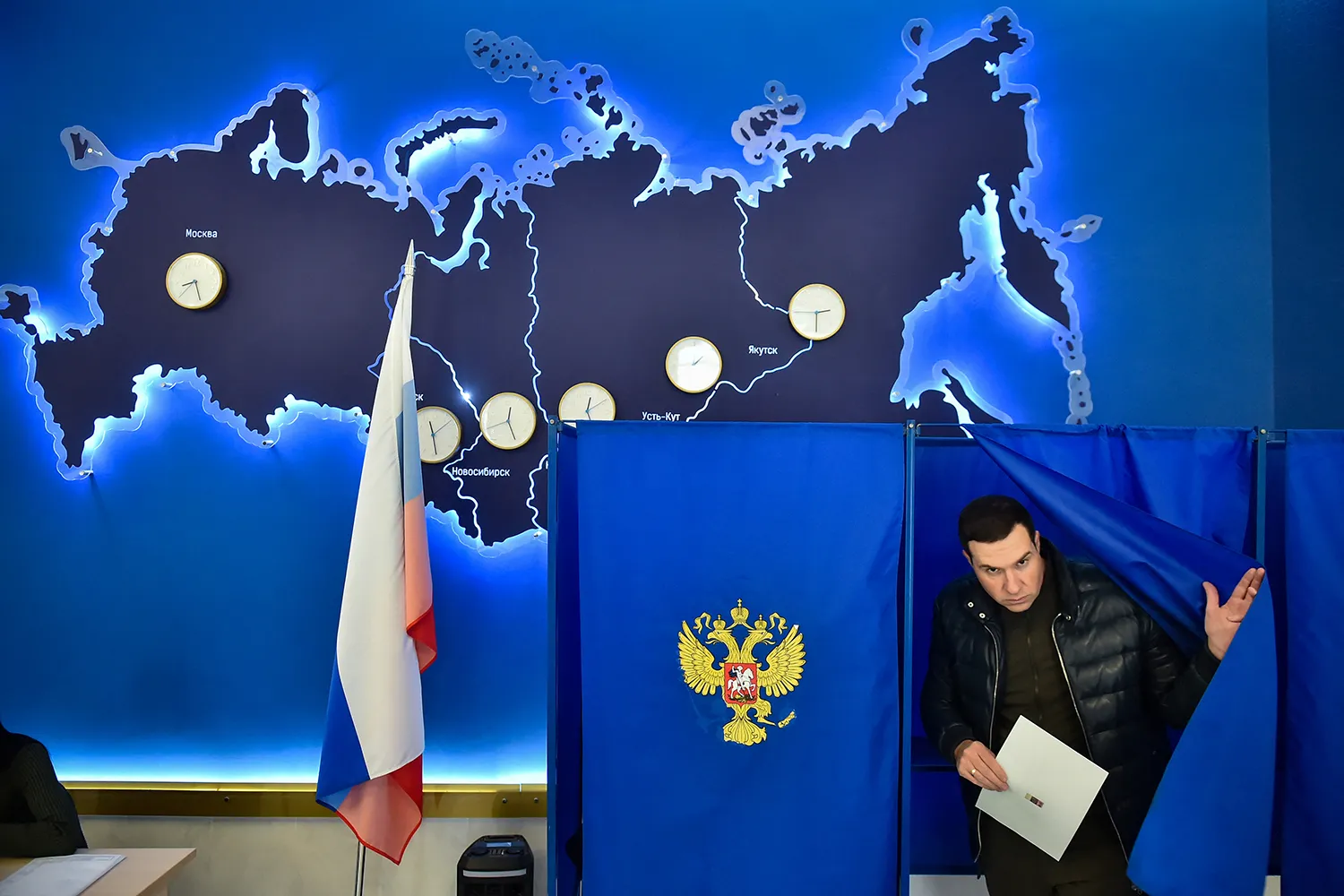 A man wearing a dark puffy coat and holding a piece of paper pushes a curtain aside as he steps out of a voting booth adorned with the crest of Russia. Behind him is a map of Russia with clocks of different time zones. A Russian flag on a stand is in front of the map.