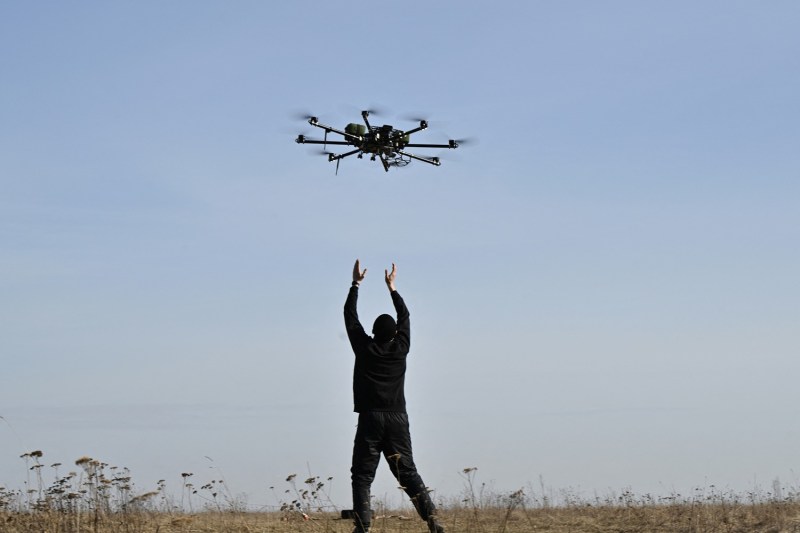 A man seen from behind stands on grass with his arms outstretched to the sky. Above his head on a blue sky is a hovering drone.