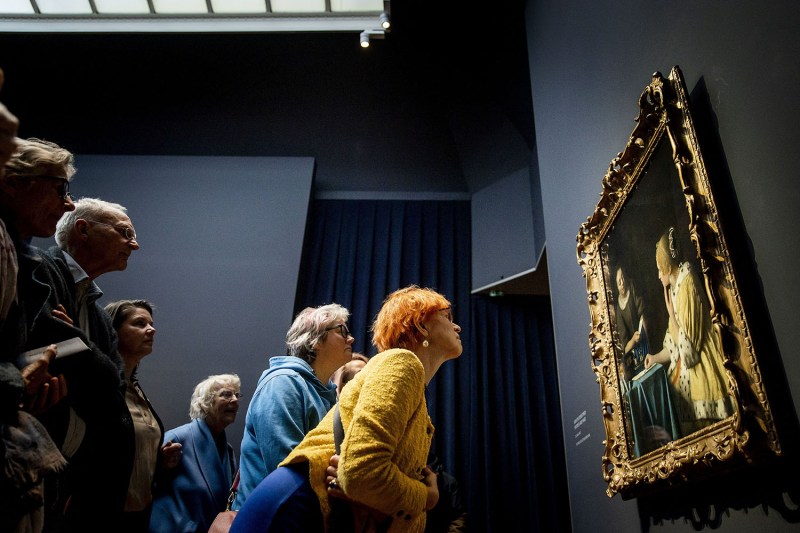 A crowd of visitors are shown from the side as they lean in to look at the painting "Mistress and Maid" by Johannes Vermeer at an art museum in Amsterdam. The painting is a portrait of two women and hangs in a gilded gold frame.