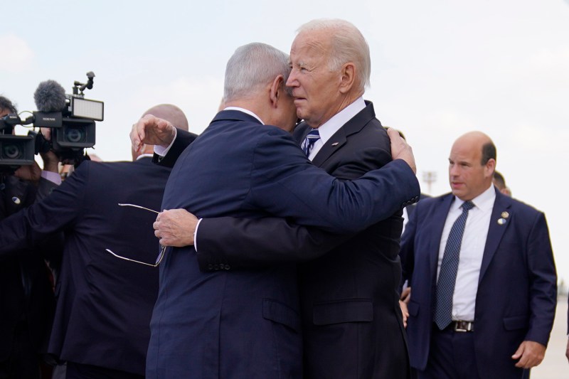 U.S. President Joe Biden, wearing a dark suit and holding sunglasses, embraces Israeli Prime Minister Benjamin Netanyahu. Photographers and other people are seen around them.