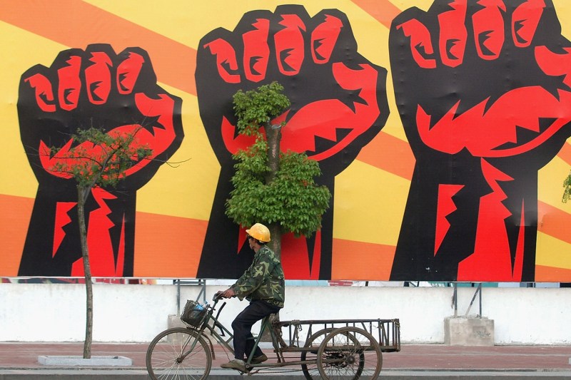 A migrant worker wearing a camouflage patterned jacket and a yellow hard hat rides a tricycle on a road in front of a building wall with a large mural. The man's head is tilted toward the mural, which shows three red and black fists against an orange and yellow striped background.