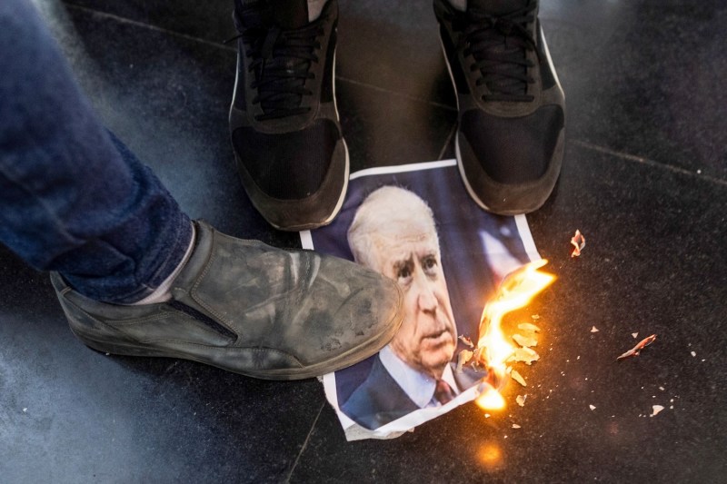 Three sneakered feet on a black tiled floor stomp on a photo of U.S. President Joe Biden. The picture's corner is alight.