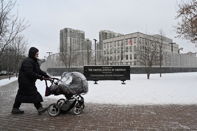 A woman bundled in a shin-length winter coat, hat, and gloves pushes a stroller past the U.S. Embassy in Kyiv. The ground is covered with snow, and the large gray complex of buildings looms against a cloudy sky.