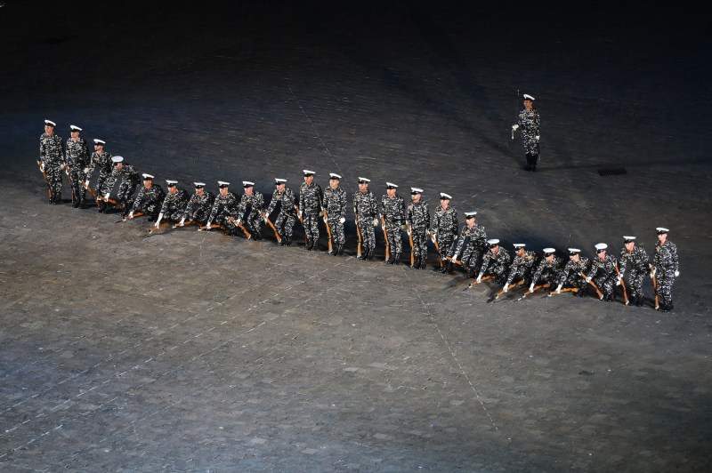 About two dozen Indian Navy sailors stand in a row on a concrete or stone floor, dressed in camouflage uniforms and white hats and holding rifles. The soldiers at the ends and center of the line stand up straight while the ones between them kneel to the ground as they rehearse a demonstration, creating the appearance of a wave or ripple down the line.