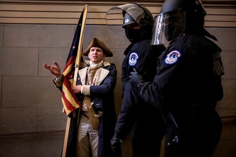 A man wearing a tricorner hat and revolutionary war uniform and holding a flag gestures as two police officers wearing riot gear pass.