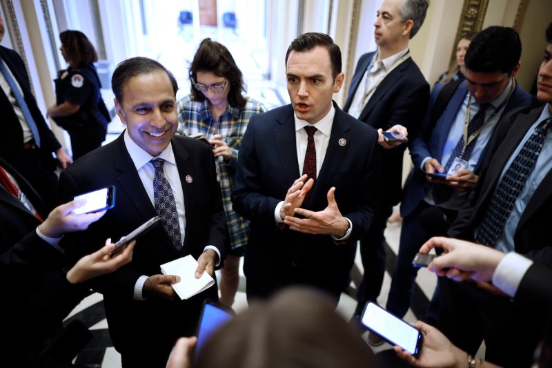 U.S. Reps. Raja Krishnamoorthi and Mike Gallager stand next to each other as they speak at the center of a cluster of reporters holding up microphones and cell phones to record their remarks. Both men wear dark suits; Krishnamoorthi is smiling, and Gallagher gestures with both hands as he speaks.