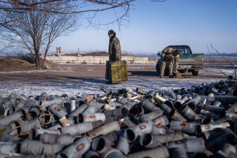 A helicopter crew member of the Ukrainian Army carries a box of ammunition in each hand as he crosses a dirt path beneath a blue sky. There are patches of snow on the ground. Another solider is visible unloading more boxes from the back of a pickup truck in the background, and piles of empty, used shells are visible in the foreground.