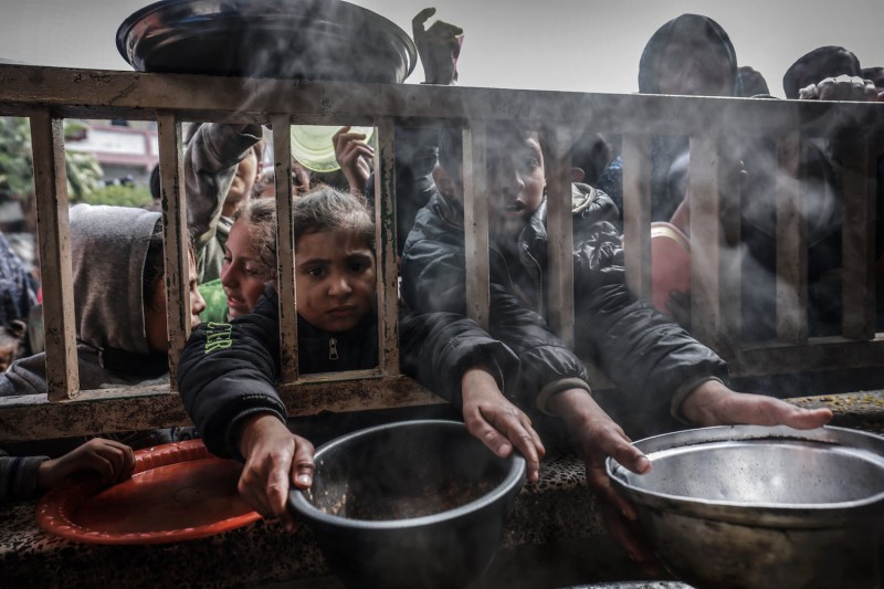 Children with dirt on their faces reach out through bars with large bowls.