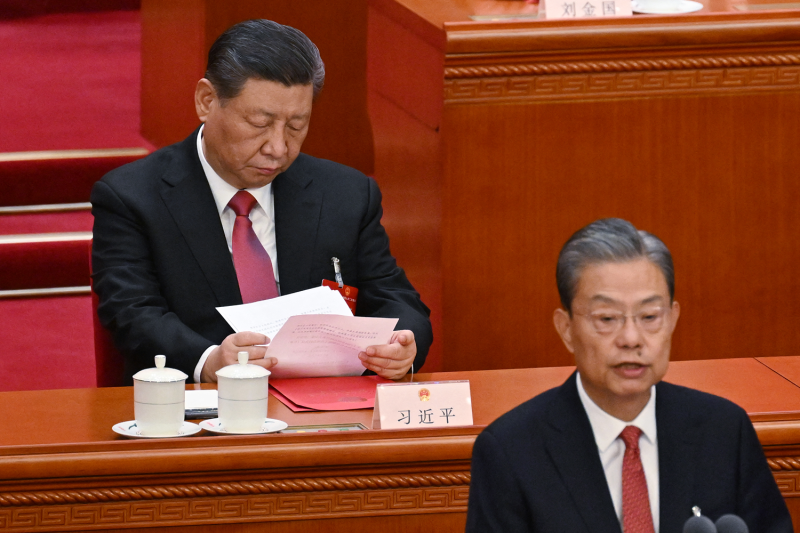 Chinese President Xi Jinping sits at a wood-paneled desk, shuffling through paper documents as he presides over a meeting of the National People's Congress in Beijing. Xi is a middle-aged man wearing a black suit and red tie, and he wears a serious expression as he reads the documents.