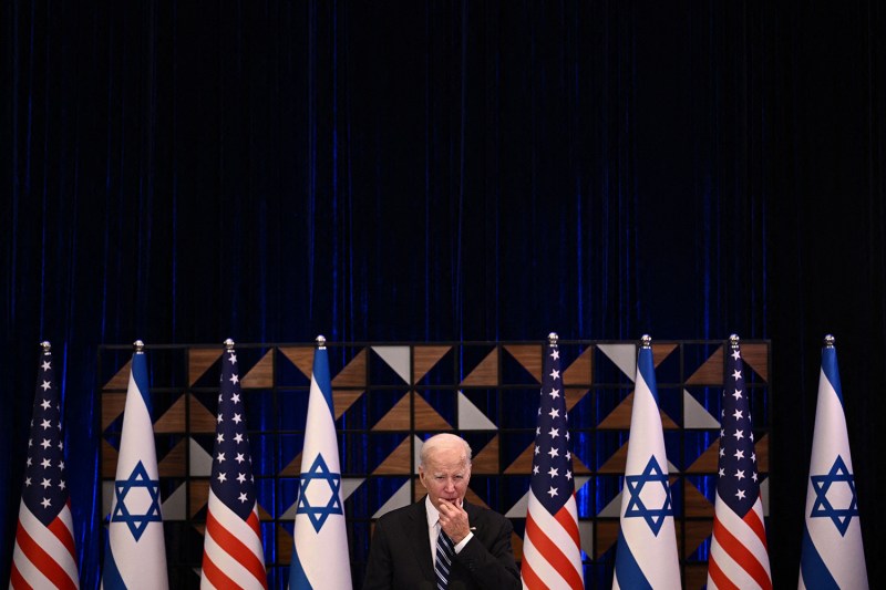 A white-haired man in a suit and tie puts his hand to his mouth and squints. He is flanked by multiple alternating flags of Israel and the United States.