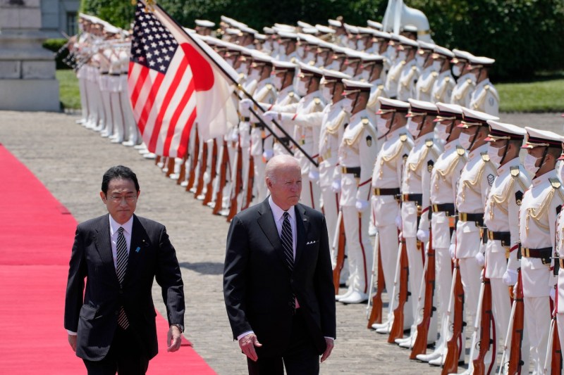 U.S. President Joe Biden and Japanese Prime Minister Fumio Kishida walk side-by-side on a red carpet as they pass by members of a Japanese honor guard wearing matching white dress uniforms and standing at attention with their rifles at their sides.