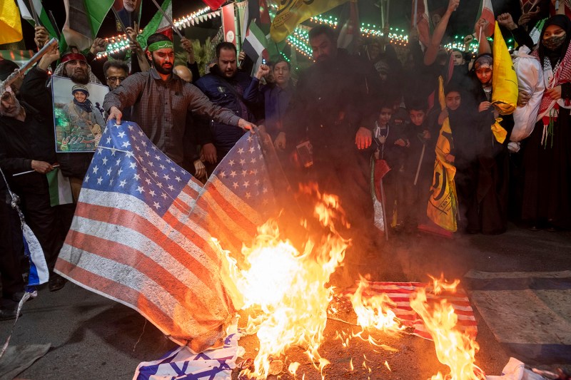 Iranian protesters stand in a crowd around a man who is feeding two large U.S. flags into a fire that already contains an Israeli flag. Protesters in teh background hod flags and chant beneath a night sky.