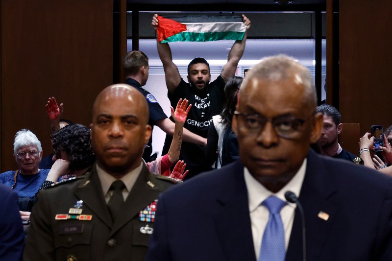 A protester holding a Palestinian flag stands up at the back of an audience of people seated in a U.S. congressional committee hearing room. Other protesters sitting around him raise up hands painted red to signify blood. In the foreground and slightly out-of-focus, Defense Secretary Lloyd Austin wears a serious expression as he sits in front of a microphone.