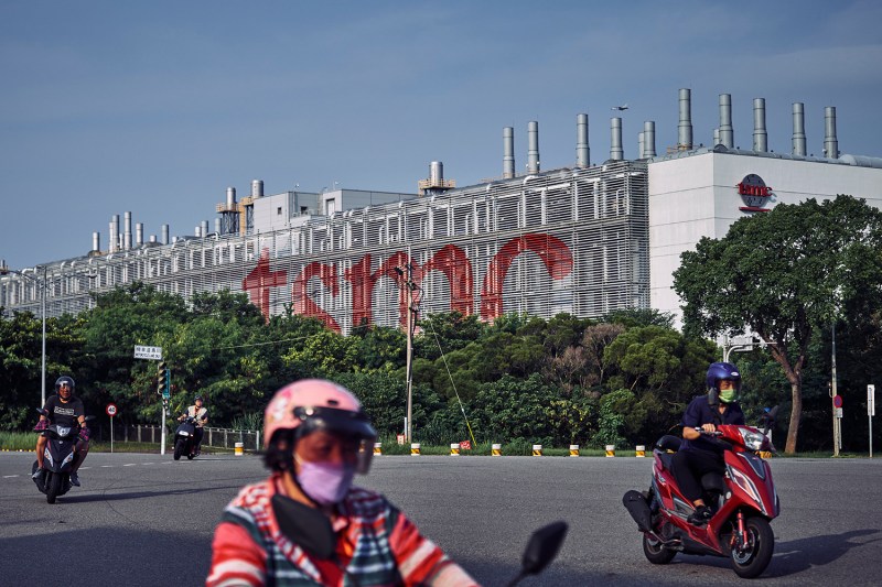 A group of people on motorbikes ride on the street in front of a massive microchip fabrication plant. The company name, TSMC, is displayed in large red letters on the front of the building.