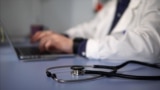 Unknown location -- A stethoscope on the table on the background of a doctor typing on a laptop