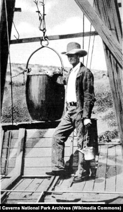 James Larkin "Jim" White standing next to a guano bucket atop the guano shaft at Carlsbad Caverns. The bucket was used to carry the first tourists into the caverns.