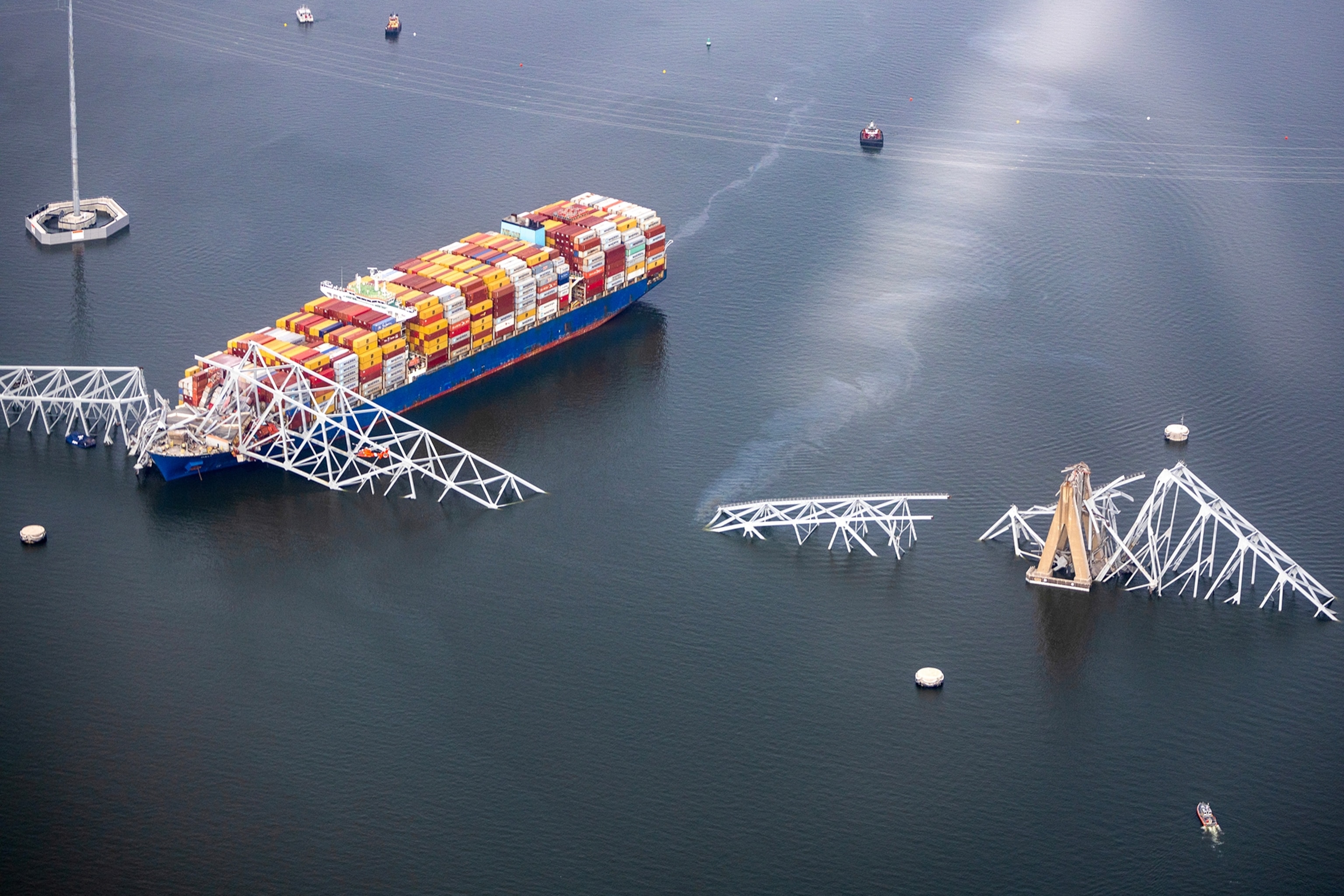 An aerial view show large broken pieces of a bridges structure poking out of the water with a contianer ship of colorful cargo.