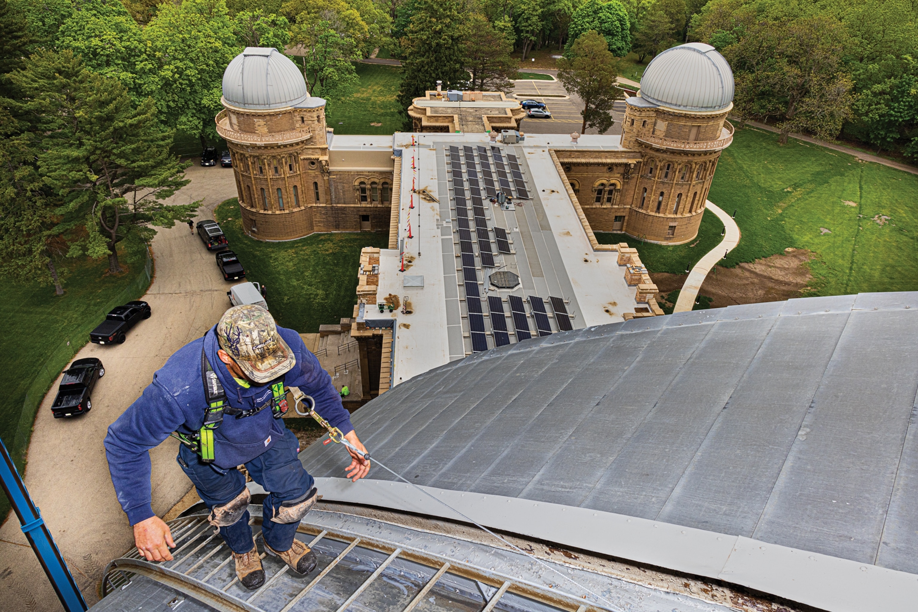 Man in hard hat climbing up the metal ladder on roof. He is attached by safety harness.