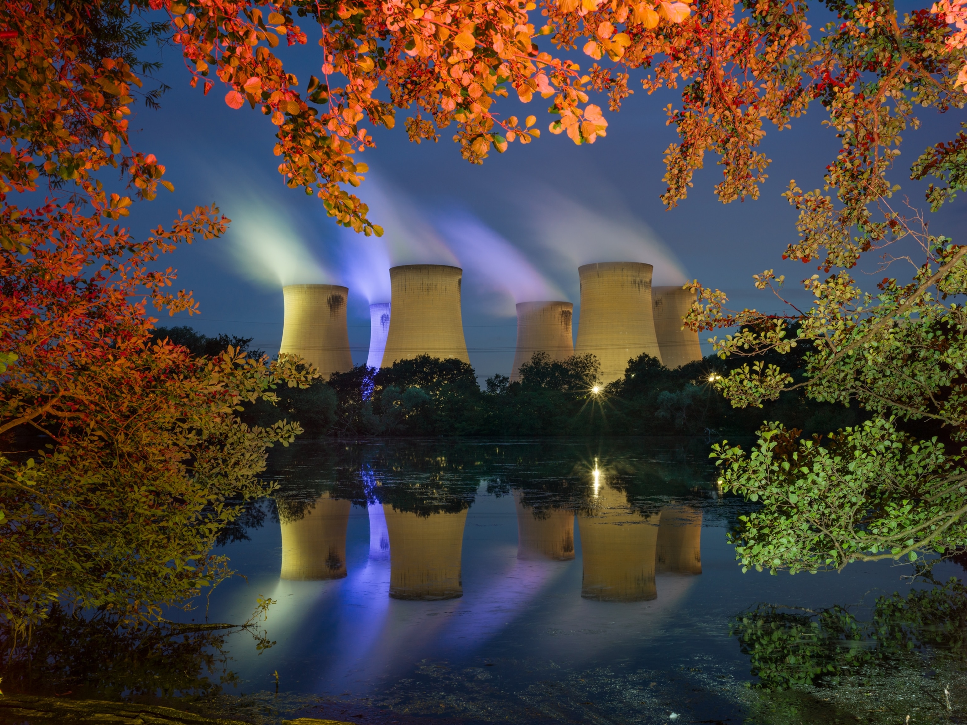 Night view of power station cooling towers from across a pond with reflection in water.