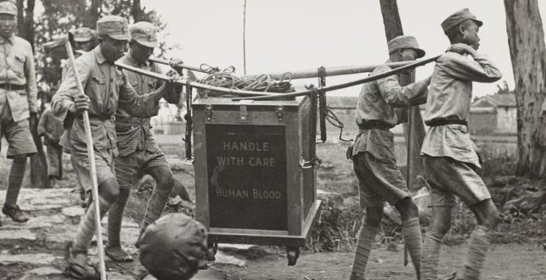 A black and white photograph of four Asian men in uniform carrying a box labeled in English "Humand Blood Handle with Care" held with poles on a dirt road.