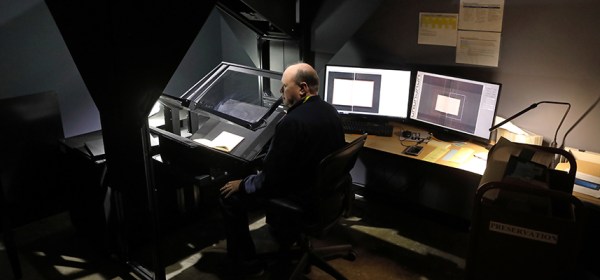 A white man sits in a darkly-lit room next to monitors showing the book he is scanning on a large format scanner
