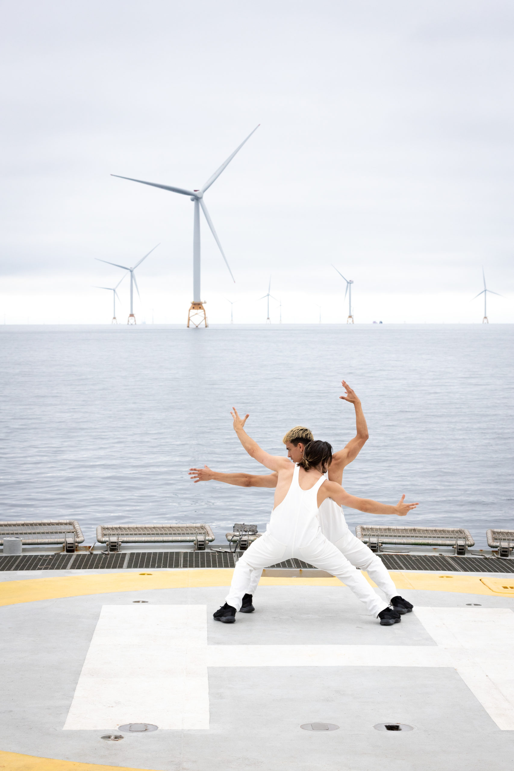 Two Dancers moving on a helicopter platform in the ocean. There are wind turbines along the horizon.