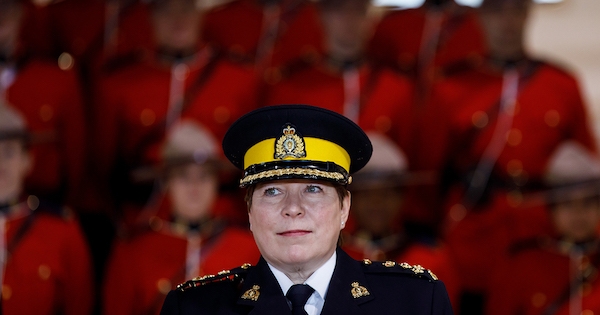A white woman wearing a black RCMP uniform, with mounties in red serge behind her
