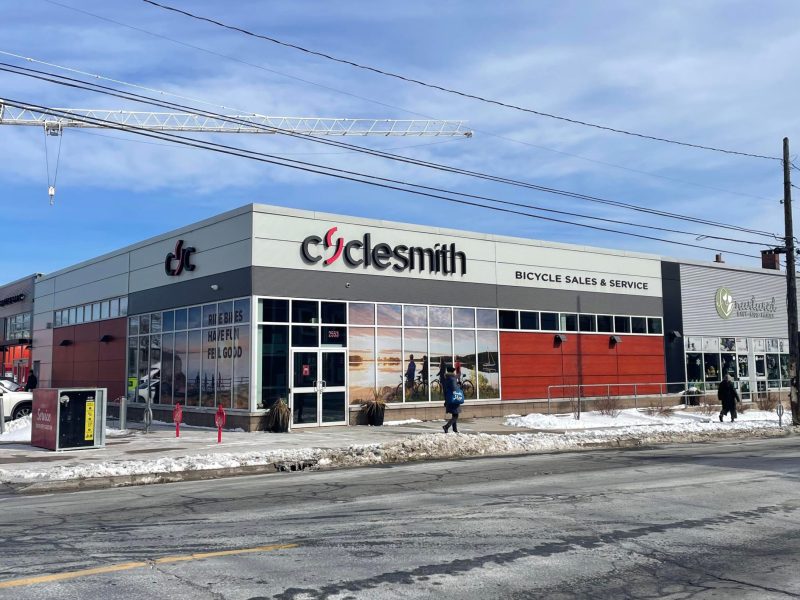 The outside of a building that has lots of windows and rust-red and grey siding. A bit of dirty snow is on the sidewalk out front and a few pedestrians walk by. The sign on the building says Cyclesmith, Bicycle Sales and Service. Hovering behind the building is a construction crane.
