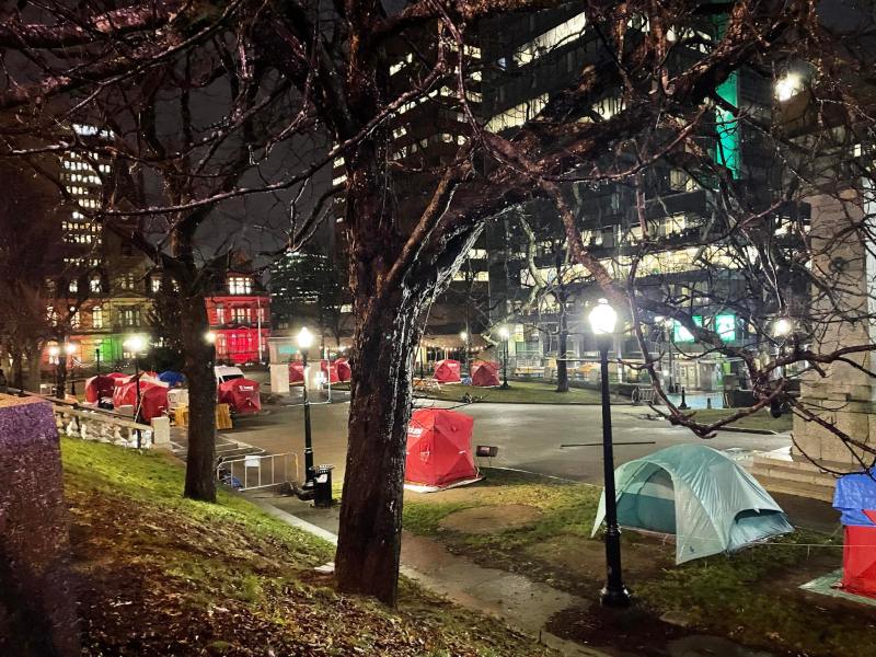 Red shelters and tents stand in the middle of a town square at night, surrounded by office buildings with floors of offices lit up.