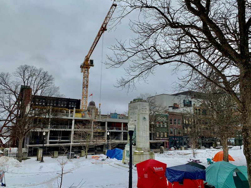 A construction crane hangs over a multi-storey building in progress and a downtown park with tents and red shelters covered in snow. A war cenotaph sits in the centre of the park.