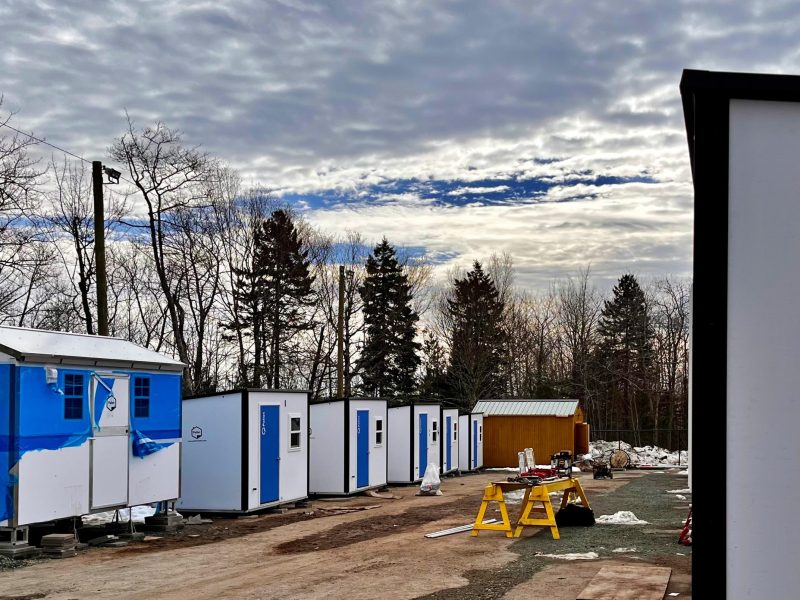 A row of white Pallet shelters with bright blue doors sit on a dirt road as a blue sky peeks out from clouds above.