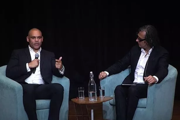 Twoi men sat in armchairs chatting on a stage in a concert hall