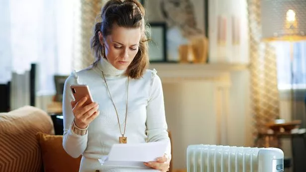 concerned young woman in white sweater and skirt sitting on couch near white electric oil radiator with smartphone and utility bill in the modern living room in sunny winter day.