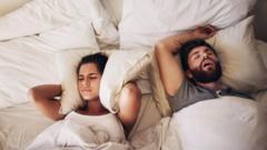 A woman covers her ears with a pillow while a man snores while sleeping