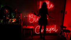 An attendee poses for a photograph inside the Little Black Chapel at The Satanic Temple's Satancon 2023. The figure is silhouetted against a wall lit by a red neon sign reading 'Little Black Chapel'. A table with candles on it stands to the left.