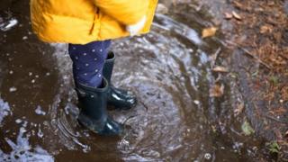 A child wearing wellington boots and a yellow rain coat standing in a puddle.