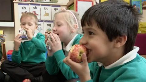 Three primary school children eating apples and drinking milk in a classroom.