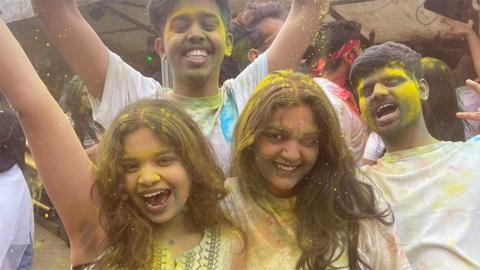 Group of four friends - two women at the front and two men at the back, celebrating the festival of Holi. There is yellow coloured powder on their faces and white shirts, and in their hair. They are shouting with their arms raised.