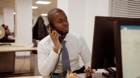 A financial industry worker speaks into his phone while looking at his computer monitor.