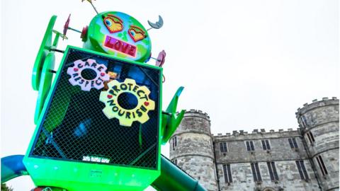 A green robot in front of Lulworth Castle