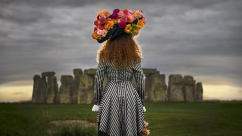 The back of a woman with long hair wearing a bright headdress with lots of pink and orange dahlias standing in front of Stonehenge with a grey sky.