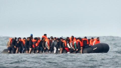 A rubber dingy on the sea, overloaded with people in orange life vests