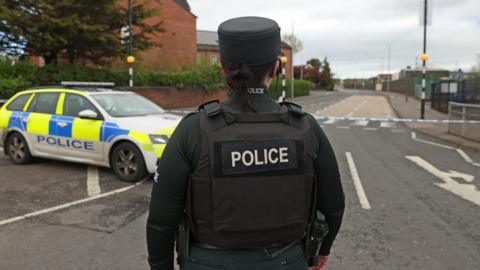 A female officer stands with her back to the camera - in front of her is a PSNI patrol car and police tape