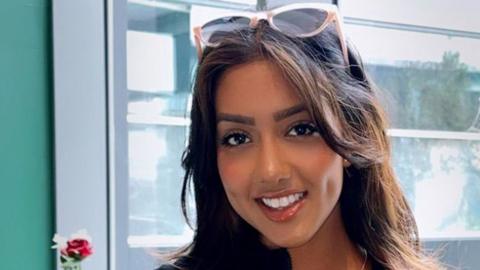 A photo of Sanam Harrinanan smiling for the camera in a cafe. She has a pair of sunglasses on the top of her head and her brunette hair is worn down. Behind her is a window.